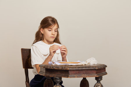 Head with threat. One adorable little girl sitting at vintage table and embroidering with excited face over light background. Concept of carefree childhood, holidays, fun, family, retro styleの写真素材