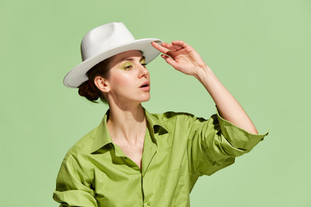New stylish clothes. Portrait with serious young woman wearing white hat and touching headdress with hand over green background. Concept of fashion, style, beauty, wellness, skincareの写真素材