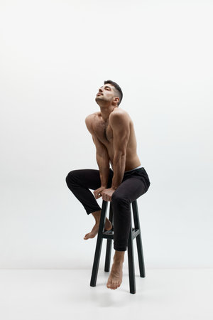 Masculinity and strength. One handsome pensive man sitting on chair, dreaming and looking up over white studio background. Concept of mens health, beauty of male body, fashion, adの写真素材