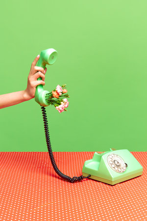 Spring season mood. Portrait with female hand holding telephone receiver with flowers inside near festive table on green studio background. Concept of nature, bloom season, beauty, art, creativity, adの写真素材