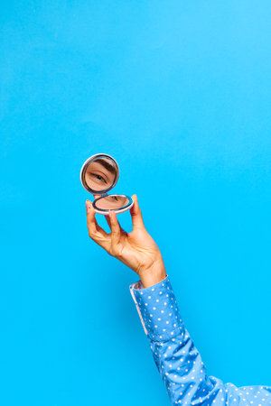 Graceful female hand holding small mirror with reflection of beautiful girls eye glowing with joy over bright blue background. Fashion, beauty, creativity, ad. Human emotions, expression conceptの写真素材