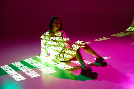 Error, mistake. Portrait of dreaming girl sitting on floor with digital neon filter lights with inscryption on body on pink mode background. Concept of digital art, fashion, cyberpunk, futurismの写真素材