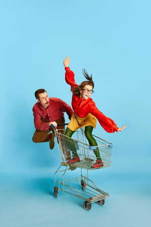 Black Friday sales. Portrait with funny couple, handsome man and excited woman riding in shopping cart over blue studio background. Concept of fun, human emotions, love, relationship, adventures, adの写真素材