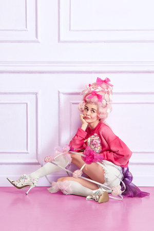 Portrait of young girl, sad princess wearing elegant pink clothes, and peruke holding candies over...
