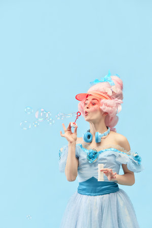 Portrait with princess, queen wearing dress playing with soap bubbles and smiling over blue studio background. Concept of comparison of eras, modernity, baroque style, beauty, fashion, human emotionの写真素材