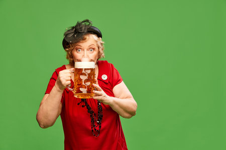Fresh beer draft. Portrait with lady, woman wearing vintage red dress holding big beer mug and drinking over green background. Concept of drinks, alcohol, degustation, festivalの写真素材