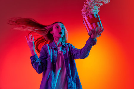 Portrait with funny, shocked, young girl, teenager in casual fashion clothes holding popcorn over red background in neon light. Concept of cinema, films, human emotions, weekends, adの写真素材
