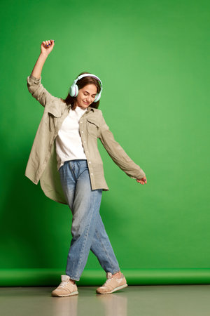 Portrait with one beautiful young girl, teenager in headphones listening to music with pleasure and dancing over green background. Concept of beauty, youth, human emotions, dreams, hobby, dance, adの写真素材