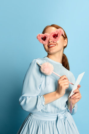 Beautiful young girl, princess with red hair wearing dress and pink plush sunglasses posing with a happy smiling face over blue studio background. Concept of medieval era, beauty, modern fashion, adの写真素材