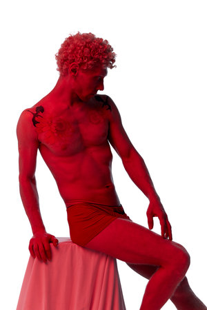 Attractive male model sitting on box covering fabric and posing shirtless in red filter isolated over white studio like statue. Concept of body aesthetics, mens beauty, creativity, inspiration, artの写真素材