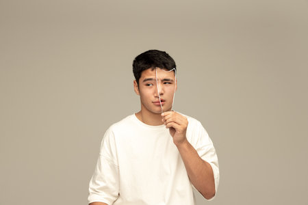 Young asian guy with calm face holding photo part of face expressing doubtful look....
