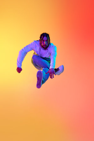 Young bearded man with dreads in casual clothes dancing breakdance against gradient orange yellow...