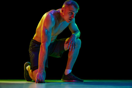 Shirtless muscular man with relief sweaty body sitting on one knee, resting after hard training against black studio background in neon light. Concept of sport, competition, health, strength, adの写真素材
