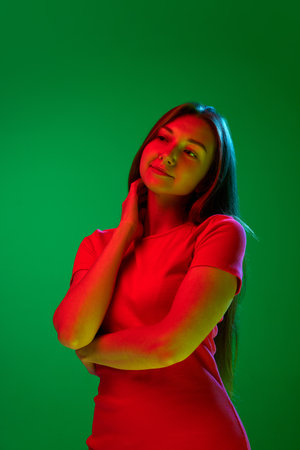 Portrait of pretty young woman in red dress tenderly posing against green background in neon light. Happiness and calmness. Concept of youth, human emotions, lifestyle, facial expression, adの写真素材