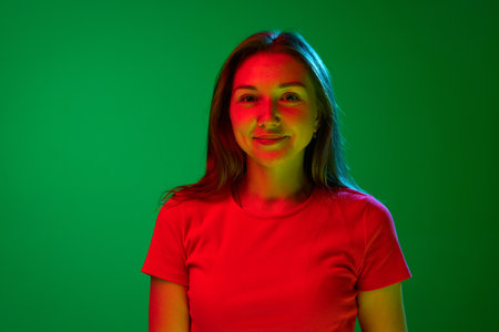 Portrait of beautiful young woman in red dress posing looking at camera with little smile against green background in neon light. Concept of youth, human emotions, lifestyle, facial expression, adの写真素材