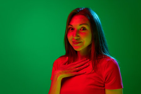 Portrait of beautiful young girl in stylish red dress posing with shocked face against green background in neon light. Concept of youth, human emotions, lifestyle, facial expression, adの写真素材