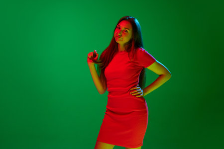 Portrait of young brunette woman in red dress standing with unpleasant face against green background in neon light. Concept of youth, human emotions, lifestyle, facial expression, adの写真素材