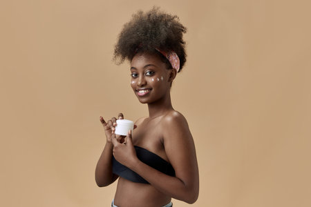 Organic cosmetics. Portrait of young, beautiful, african woman applying face moisturizing cream, spf against studio background. Concept of natural beauty, skin care, health, fashion, wellness, adの写真素材