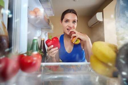 Young woman opening fridge, looking inside and choosing what to eat. Eating sausage and vegetables. View from inside of fridge. Concept of food, diet, emotions, domestic lifestyle, tasteの写真素材