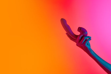 Womans hand holding vibrator, massager over yellow-pink background in neon light. Concept of toys for adult, healthy lifestyle. Copyspace for ad, textの写真素材