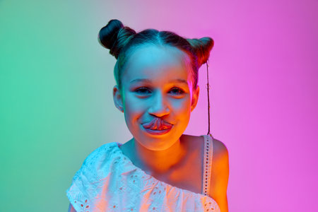 Grimace. Charming beautiful little girl, child looking at camera with smile over gradient neon light background. Concept of childhood, emotions, kids fashion. Model looks happyの写真素材