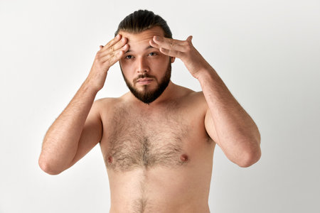 Portrait of handsome man having beard looking away and toching area under eyes over grey background. Concept of beauty, self care, cosmetic, procedures, health, natural, ad.の写真素材