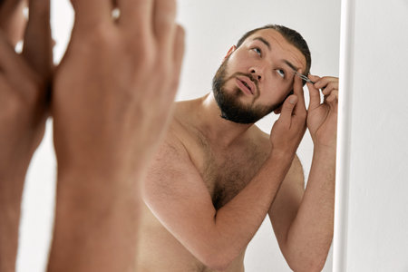 Photoshoot of man looking in mirror and shaping with tweezers over isolated grey background. Concept of beauty, appearance, self care, procedures, health, natural, ad.の写真素材