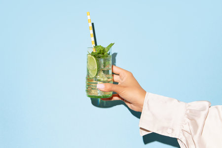 Ladys hand holding glass of mojito with lime on light blue background. Concept of copy space for ad, design, party, relax, pop art.の写真素材