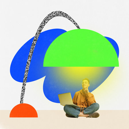 Young boy sitting on floor under big drawing lamp and doing homework through laptop over blue and white background. Concept of education, youth, study, college lifestiyle, graduation and ad.の写真素材