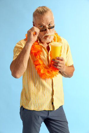 Middle-aged grey-head man, stylishly, brightly dressed in summer, holds a refreshing cocktail, looking into the camera, pushing his glasses away. Concept of emotion, rest, recreation, holidays. Ad.の写真素材