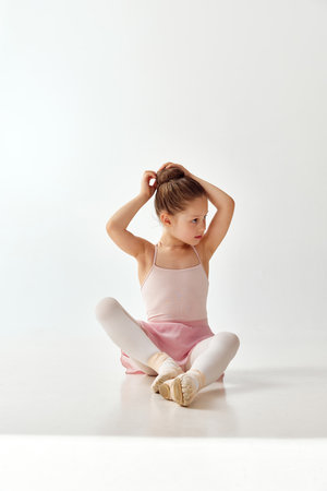 Portrait of small adorable ballerina dancer girl correct her hair bundle in rose tutu ballet dress, in pointe, white legging against on white studio background. Concept of beauty, fashion, hobby. Adの写真素材