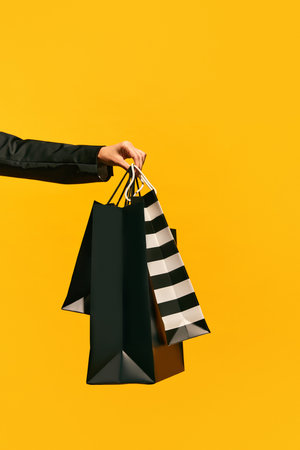 Sale offer. Black Friday. Shopping delivery. Closeup of ladys hand carrying purchase black bags isolated on bright yellow background. concept of fashion, beauty, salesperson. copy space. adの写真素材