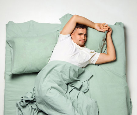 Young attractive man lying looking to camera, covered with blanket raising up stretching arms in wake up. Enough night sleep. early morning. Concept of recreation, rest, sleep wellness, home.の写真素材
