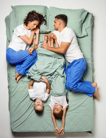 Top view portrait of people covered with blanket lying in bed. Children lying crossed bed and parents on edge of bed. Family sleeping. Enjoying being together. Concept of sleep wellness, family time.の写真素材