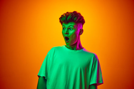 Portrait of surprised, shocked young man with open mouth in white t-shirt against gradient orange background illuminated neon light. Concept of youth, beauty, fashion, trends, emotions, sale.の写真素材