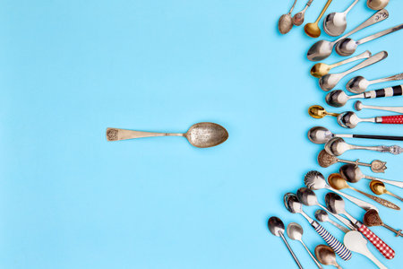 Tips of cutlery. Top view photo one table spoon surrounded of variety of antique silverware and gold spoons against blue studio background. Concept of food, holiday, table setting, retro, vintageの写真素材