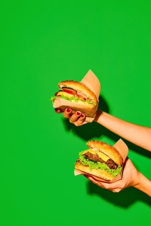 Delicious fast-food. Hands holds appetizing hot hamburger and cheeseburger in paper against vivid green background. Concept of junk food, menu, delivery, catering, take away. Copy space. Adの写真素材