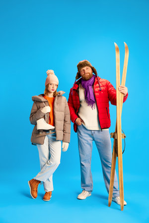 Full length portrait of father and daughter dressed winter warm outfit and holds vintage ski and skates isolated blue studio background. Concept o winter, extreme sport, hobby, trip, relax, parenthoodの写真素材