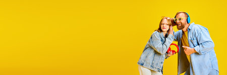 Banner. Father and daughter, parent and kid dressed denim outfit and have fun listening music from one headphones over yellow background. Concept of parenthood, childhood, family, style. Copy space.の写真素材