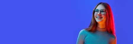 Banner. Portrait of young girl, teenager with beautiful smile in style glasses isolated blue background in mixed neon light with negative space to insert text. Concept of beauty, fashion and style. Adの写真素材
