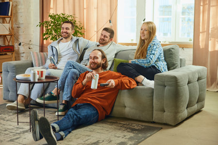 Photo of happy cheerful people, friends sitting in living room and watching comedy film, TV show. Concept of movie time, friendship, togetherness, TV program, online event, recreation, relaxation.の写真素材