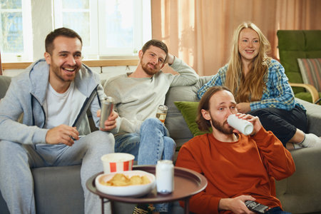 Group portrait. Guys and girl sitting in living room and spending all time watching comedy series and having fun. Concept of friendship, togetherness, TV program, online event, recreation.の写真素材