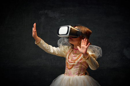 Portrait of medieval person, little princess playing games in modern VR glasses against dark vintage background. Concept of historical remake, comparison of eras, medieval fashion, emotions.の写真素材
