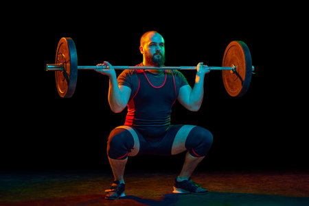 Muscular, strong young man, bodybuilder doing squats and lifting barbell, heavy weights over black background in neon. Concept of sport, strength, gym, healthy lifestyle, power, weightliftingの写真素材