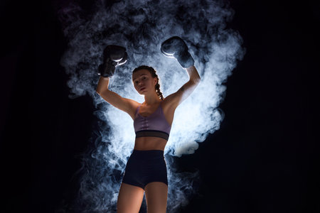 Powerful boxer girl in shorts and gloves vining pose, symbolizing strength and competitiveness in sports against black background in stage smoke. Sport, active lifestyle, health, competition concept.の写真素材