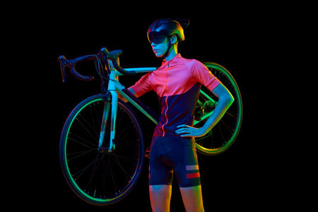 Young man in sportswear, helmet and googles holding bicycle on shoulder isolated over black studio background in neon light. Concept of sport, action, competition, power and endurance, healthの写真素材