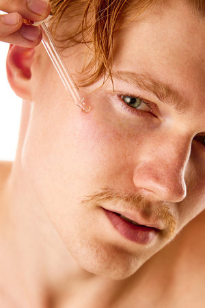 Close up, cropped portrait of redheaded man with mustache applying serum, oil from pipette on face against white studio background. Concept of natural beauty, selfcare, skincare, hygiene, fashion. Ad.の写真素材
