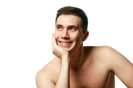 Attractive man, well-groomed guy with bare shoulders smiling and put his head on his hand against white background. Concept of beauty treatment and hygiene, body care, anti-aging procedures.の写真素材