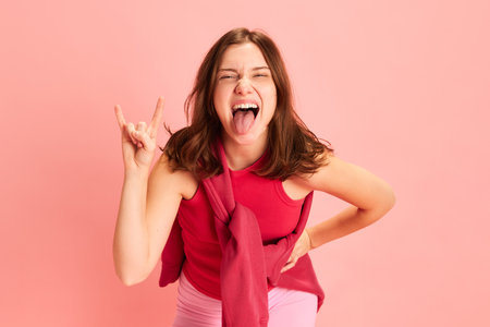 Portrait of young expressive, emotional brunette lady dressed sportwear and show rock gesture against pink studio background. Concept of sport, active lifestyle, fitness, music, hobby, energy.の写真素材