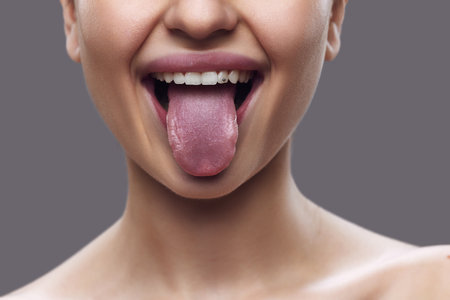 Cropped photo. Close-up of female mouth with tongue, highlighted by subtle lip gloss, against gray background. Concept of techniques to improve articulation and muscle control in speech.の写真素材